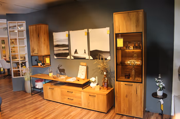 Wohnzimmerausstellung mit modernen Möbeln aus Massivholz in Eiche, bestehend aus Sideboard, Vitrine und Hängeschrank vor einer dunkelgrauen Wand mit dekorativer Beleuchtung und Wandbildern.